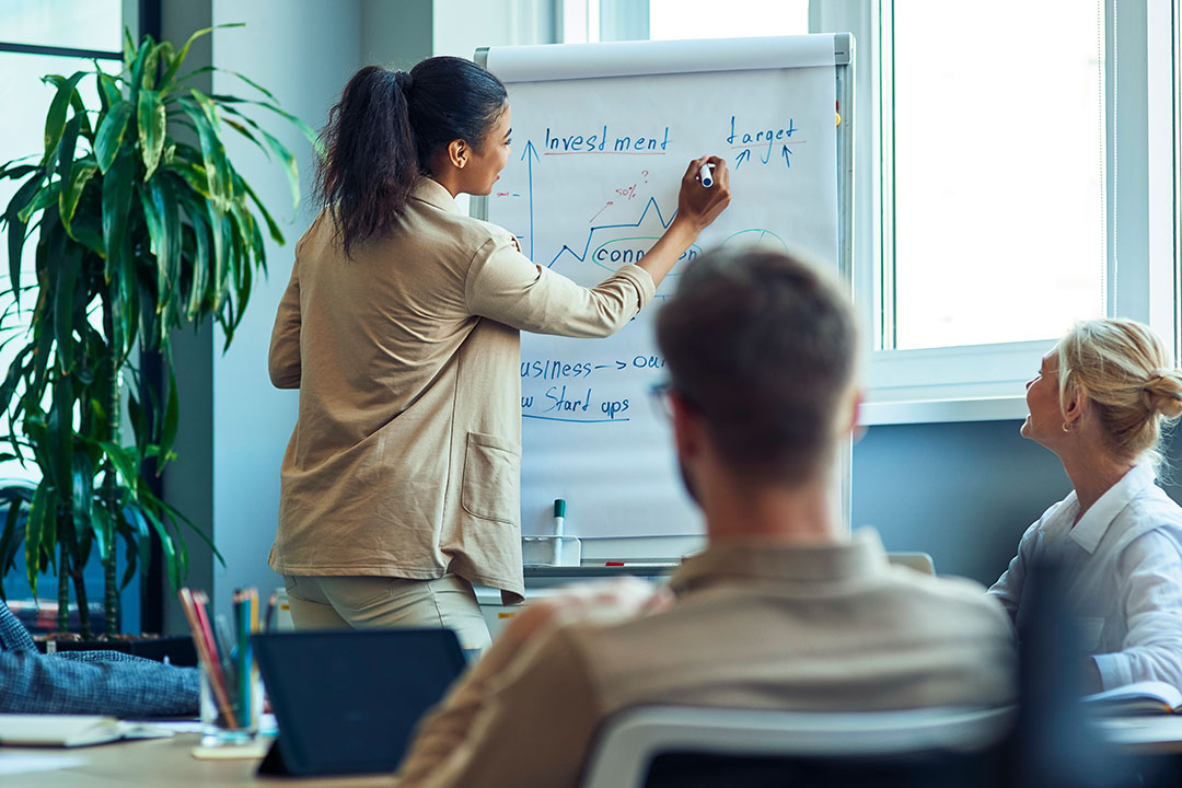 Referentin erklärt am Flipchart Investment und Zielsetzung, zwei Teilnehmende hören zu: Weiterbildung im Unternehmen